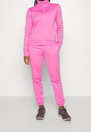 Osoba ubrana w jasnoróżową rozpinaną bluzę sportową i pasujące spodnie dresowe oraz ciemnoszare sneakersy, stojąca na tle jednolitego jasnego tła.