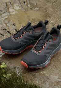 Schwarz-orangefarbene Adidas Terrex Trailrunning-Schuhe auf matschigem Boden mit Pfützen und grünen Gras in der Nähe.