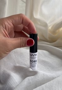 Bâton de remplissage des pores NYX Professional Makeup dans un tube blanc avec des accents noirs. Texturé lisse, de forme cylindrique, tenu par une main avec du vernis à ongles rouge.