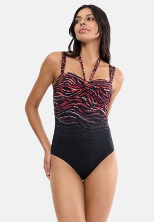 Vrouw die een zwart-rood dierenprint badpak met halter- en schouderbanden draagt, staand tegen een effen lichte achtergrond.