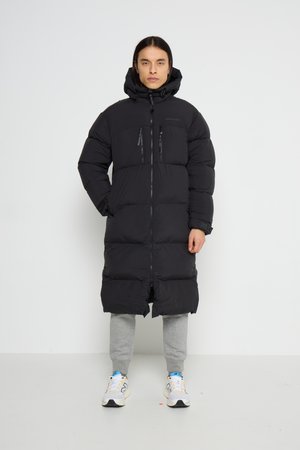 Person mit langen dunklen Haaren, die einen schwarzen langen Steppmantel mit Kapuze, graue Jogginghosen und weiße Turnschuhe trägt, vor einem einfarbigen weißen Hintergrund stehend.