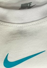 Maglietta Nike bianca con colletto corto, che presenta un grande logo swoosh Nike in teal. Tessuto texture con dettagli di cucitura visibili.