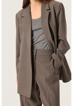 Kobieta w taupe dopasowanej marynarce i pasujących do niej spodniach z wysokim stanem, na szarym, gładkim t-shircie, z jedną ręką w kieszeni.