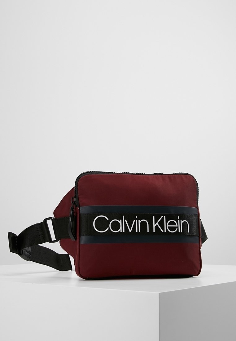 Bordeaux kleurige schoudertas van stof met een zwarte riem, voorzien van het logo "Calvin Klein" in het wit. Ritssluiting en verstelbare riem voor gebruiksgemak.