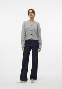 Cardigan en maille gris avec un col rond, des boutons sur le devant et des manches longues. Porté avec un jean large bleu foncé et des chaussures couleur crème.