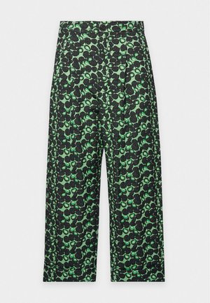 Un mannequin se tient devant un fond de studio uni, vêtu d’une chemise blanche sur un haut noir, d’un pantalon court vert à motifs et de sandales noires à talons.
