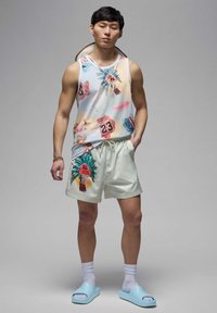 Uomo che indossa una camicia senza maniche con stampa tropicale e pantaloni corti coordinati, calzini bianchi e ciabatte blu, in piedi con una mano in tasca.
