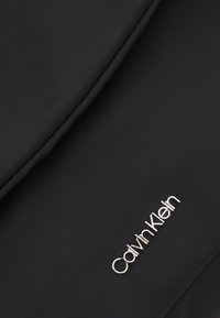 Tissu noir avec un logo Calvin Klein argenté en relief dans le coin inférieur droit, montrant une texture détaillée et des coutures.