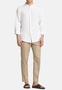 Camicia bianca in lino con colletto alla mandarina, abbinata a pantaloni beige e sandali marrone scuro. Abbigliamento leggero e informale.