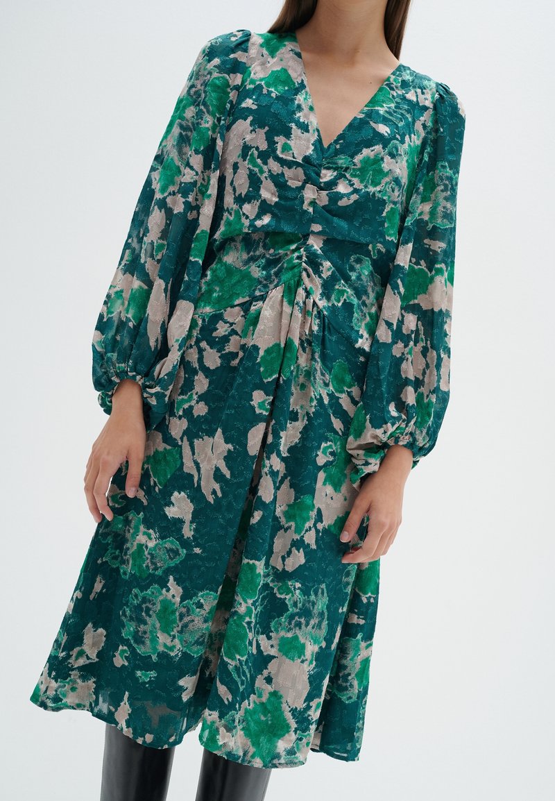 Robe florale verte avec un décolleté en V, de longues manches bouffantes et un détail de taille froncée. Elle présente un tissu léger avec une texture douce.