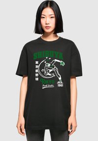 Sort bomulds T-shirt med et stort grafisk print med "SHIBUYA" i grøn og hvid, som viser en skater og tekstdetaljer, afslappet pasform.