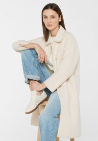 Beige Übergröße-Mantel mit Seitentaschen, getragen über einem weißen Oberteil. Hellblaue, ausgefranste Jeans und weiße Sneakers mit Kautschuksohle.