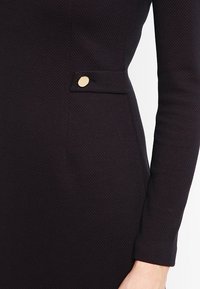 Svart klänning med ett texturerat tyg, som har en slim fit, långa ärmar och en guldknytt accent på vänster höftficka.