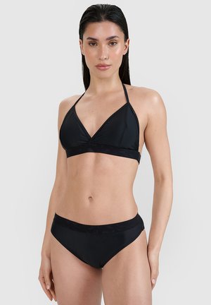 Kobieta o długich ciemnych włosach, ubrana w czarny halter bikini top i pasujące do niego dół bikini na jednolitym jasnym tle.