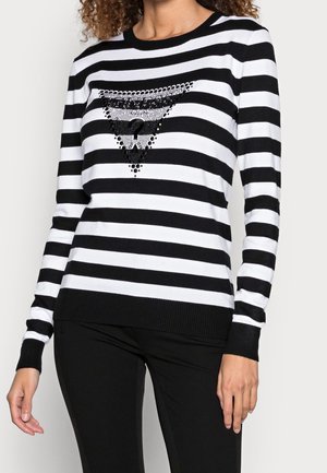 Femme portant un haut à manches longues rayé horizontalement noir et blanc avec un motif triangulaire clouté noir sur la poitrine.