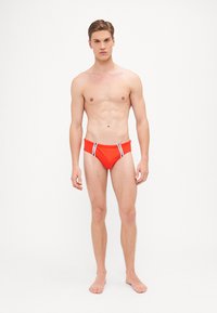 Rote Schwimmslips mit kontrastierenden blauen und weißen Seitenstreifen, hergestellt aus glattem, dehnbarem Material. Enganliegendes Design, mittlere Bundhöhe.