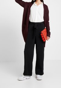 Cardigan largo burdeos, camisa blanca de botones, pantalones anchos negros y un bolso de mano con pompón naranja brillante. Zapatillas deportivas blancas.