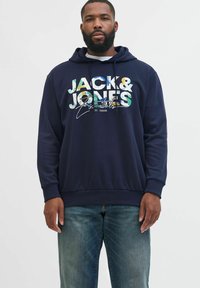 Marineblå hoodie med "JACK & JONES Essentials" i multicolor tekst, med en hvid indvendig foring og ribbede manchetter og kant; båret med jeans.