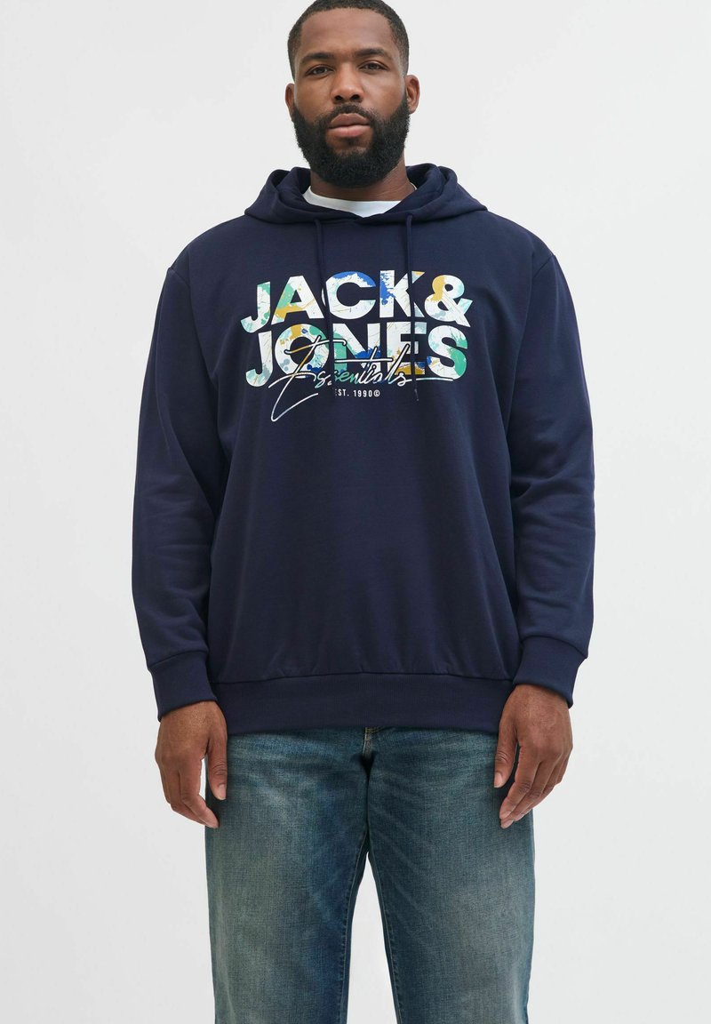 Marineblå hoodie med "JACK & JONES Essentials" i multicolor tekst, med en hvid indvendig foring og ribbede manchetter og kant; båret med jeans.