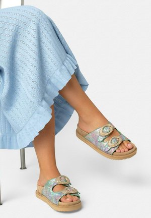 Pies con sandalias tipo slide en verde pastel y rosa, con hebillas grandes trenzadas, combinadas con una falda de punto azul claro con dobladillo con volantes.