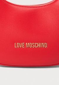 Sarkans ādas rokassomu ar zeltītu "LOVE MOSCHINO" logo, kas centrā novietots tuvu apakšai.