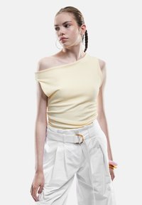 Haut jaune sans manches avec tissu texturé, associé à un pantalon plissé blanc taille haute et une ceinture avec boucle en or.