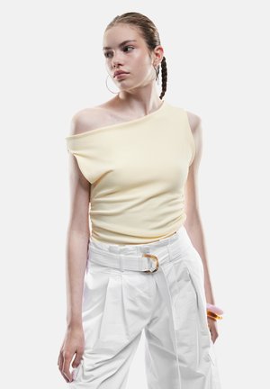 Geel off-shoulder top van textuurstof, gecombineerd met hoge witte plooibroek en een riem met gouden hardware.