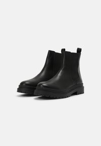 Botas Chelsea de cuero negro con paneles laterales elásticos, una lengüeta y una suela de goma gruesa y texturizada. Acabado suave y altura hasta el tobillo.