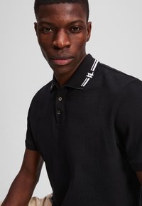 Černé polo tričko vyrobené z texturované tkaniny, s kontrastním bílým pruhem na límci a dvěma knoflíky, s detailem loga na límci.
