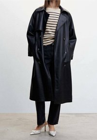 Cappotto doppiopetto in pelle nera con ampio colletto, indossato sopra un maglione beige a righe e jeans scuri, abbinato a tacchi bianchi a punta.