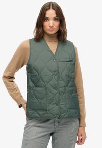 Pikowana zielona kamizelka z dekoltem w kształcie litery V, z dwiema przednimi kieszeniami i zapięciem na guzik. Noszona na brązowym swetrze z golfem i w pasiastych spodniach.