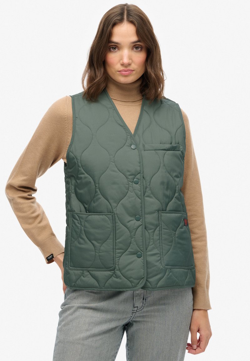 Pikowana zielona kamizelka z dekoltem w kształcie litery V, z dwiema przednimi kieszeniami i zapięciem na guzik. Noszona na brązowym swetrze z golfem i w pasiastych spodniach.