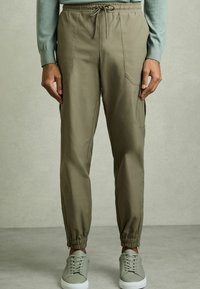 Pantaloni jogger khaki cu talie elastică, șnur și mansete înguste. Include două buzunare frontale și o finisare netedă, texturată.