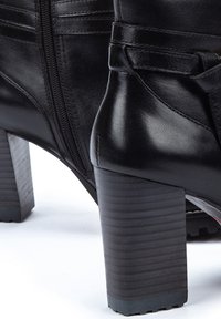 Botas de tobillo de cuero negro con un tacón grueso, que cuentan con un cierre lateral y un detalle decorativo de correa. Textura suave con un acabado pulido.