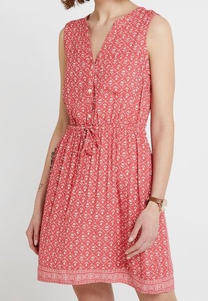 Ärmelloses korallenfarbenes Kleid mit weißem geometrischem Blumenmuster, V-Ausschnitt mit Knöpfen, Kordelzug in der Taille, getragen mit brauner Uhr am linken Handgelenk.