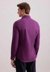 Uomo che indossa una camicia a maniche lunghe viola aderente e pantaloni scuri, in piedi di lato di fronte a uno sfondo chiaro.