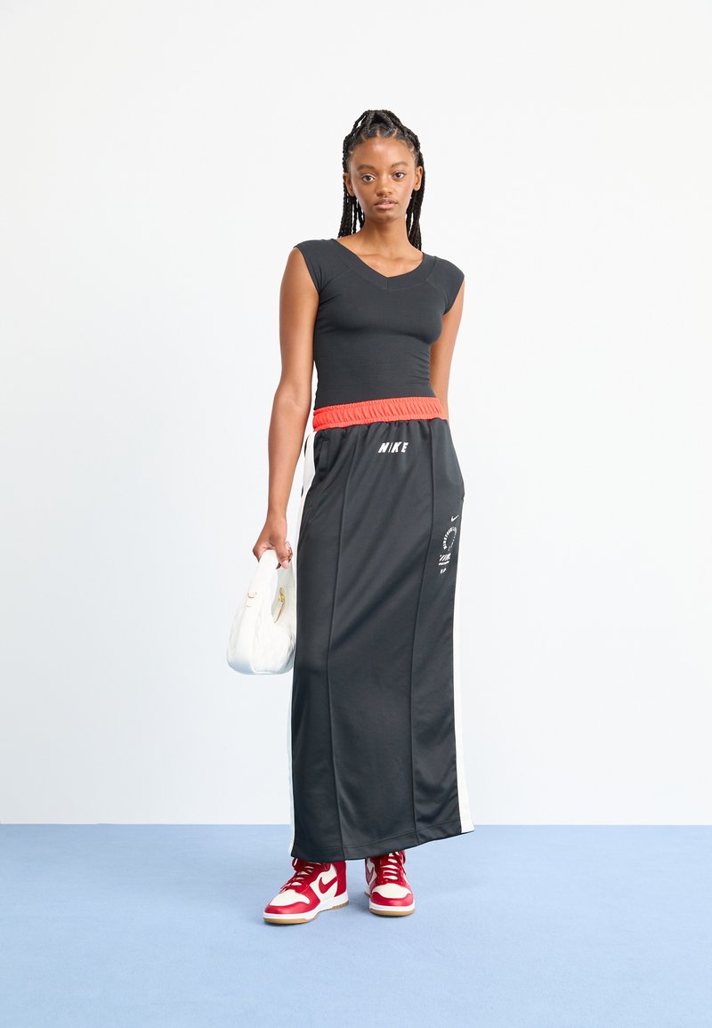 Blusa negra ajustada de mangas cortas, combinada con una larga falda negra que tiene una cintura elástica roja y franjas blancas a los lados. Zapatillas rojas.
