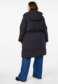 Zwarte pufferjas met capuchon, elastische taille en een middellange ontwerp. Gepaarde met wijde denim broek en zwarte schoenen.