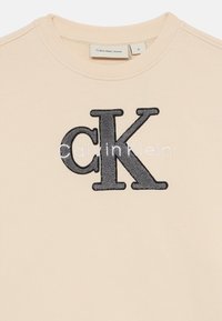 Sudadera beige con cuello redondo acanalado. Presenta un gran logotipo bordado en negro "CK" centrado en la parte delantera con el nombre de la marca en blanco debajo.
