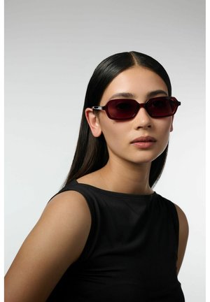 Femme aux longs cheveux foncés portant des lunettes de soleil rectangulaires rouge foncé et un haut noir sans manches sur un fond clair uni.