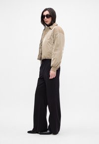 Light tan, shiny bomber jacket with a collared neck, worn over a black long-sleeve top. Paired with wide, black pinstripe trousers and sleek shoes.