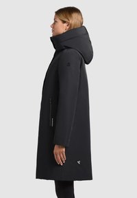 Sort polstret jakke med høj krave og påsat hætte. Har sidelommer og et logo-patch nær kanten. Strømlinet design, glat tekstur.