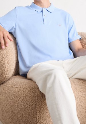 Man wearing light blue polo shirt and white pants sitting on beige textured armchair with arm rested on chair.