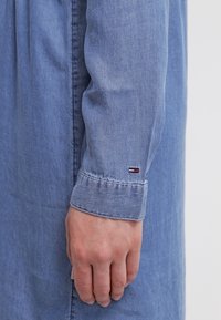 Světle modrá denímová košile s knoflíkovým manžetovým rukávem a malým logem v červené, bílé a modré barvě na rukávu. Hladká textura, uvolněný střih.