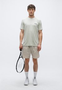 Svetlo siva majica, ki odvaja vlago, z napisom "BOSS", kombinirana z bež kratkimi hlačami. Model drži črno-belo teniško lopar.