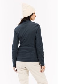 Una donna che indossa una giacca in pile blu scuro, pantaloni crema e un berretto di maglia crema, girata di spalle contro uno sfondo bianco semplice.