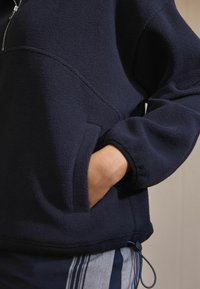 Marinblå fleece-pullov med kvartsdragkedja, avslappnad passform, framtaske och justerbar kantdetalj. Mjuk textur och minimalistisk design.