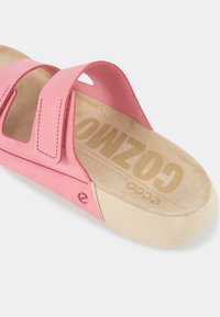 Rožinė šlepetė su reguliuojamais Velcro dirželiais, su suknele panašiu viršumi, paminkštintu padu ir kreminės spalvos padais. Papuošta iškilniu logotipu.