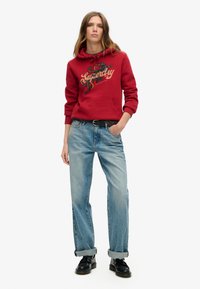 Sudadera roja con estampado gráfico, bolsillo frontal, combinada con vaqueros anchos de color azul claro, con puños doblados, y zapatos negros. Fondo blanco liso.