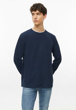 Junger Mann mit lockigem Haar, der ein marineblaues Langarmshirt und blaue Jeans trägt, steht mit den Händen hinter dem Rücken und schaut zur Seite.
